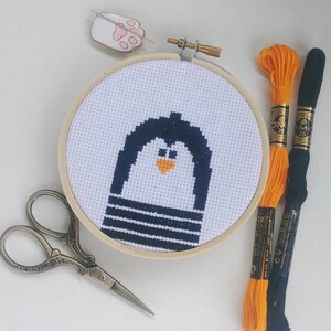 Könnte beinhalten: Ein Kreuzstich-Stickrahmen mit einem Pinguin-Design in Marineblau und Orange. Bastelbedarf: orangefarbenes und schwarzes Stickgarn, kleine Schere und ein Katzenpfoten-Nadelkissen. Der Rahmen hat einen Durchmesser von etwa 10 cm.