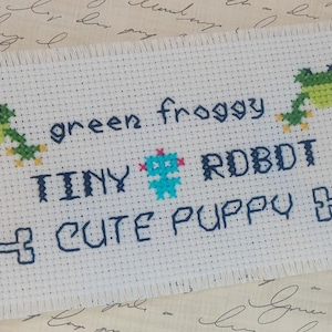 May include: A white cross-stitch fabric with the words "green froggy", "TINY RDBDT", and "CUTE PUPPY" in dark blue thread. The design includes green frogs, a blue robot, and dumbbell shapes.