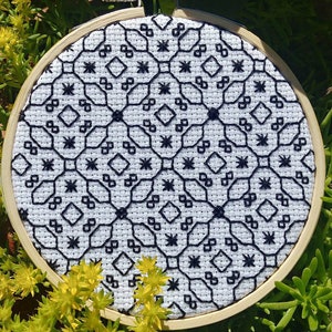 May include: A cross-stitch embroidery with a geometric black pattern on white fabric. The design features diamond and star shapes. The embroidery is displayed in a round wooden hoop, suitable for wall decoration.
