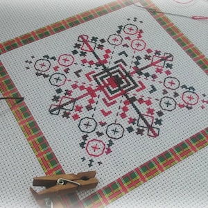 Op de afbeelding: Een voltooid kruissteekproject met een geometrisch ontwerp in zwart en rood op witte stof. Het stuk is omlijst met een rood, groen en geel geruite rand. Naai-attributen, waaronder een schaar, draad en een naald, zijn zichtbaar.