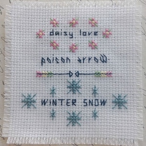 May include: White fabric with cross-stitch embroidery. The top row features pink flowers with the text "daisy love". The middle row features a green, pink, and yellow arrow with the text "poison arrow". The bottom row features blue snowflakes with the text "winter snow".
