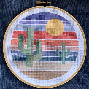 May include: A cross-stitch embroidery hoop featuring a desert landscape. The design includes a yellow sun, green cacti, and a striped sunset in shades of orange, red, purple, and blue. The hoop is made of wood and is round.
