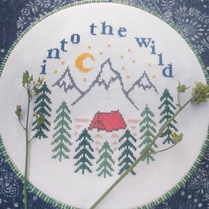 May include: A cross-stitch embroidery hoop with a camping scene and the text "Into the Wild." The design includes mountains, a crescent moon, stars, a red tent, and green trees. The hoop has a green and white border.