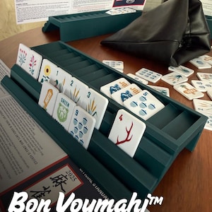 May include: A dark green plastic Mah Jongg rack holding white tiles with various nature-themed illustrations, numbers, and text. A black leather pouch, a printed rule sheet, and additional loose tiles are arranged around the rack on a wooden surface.