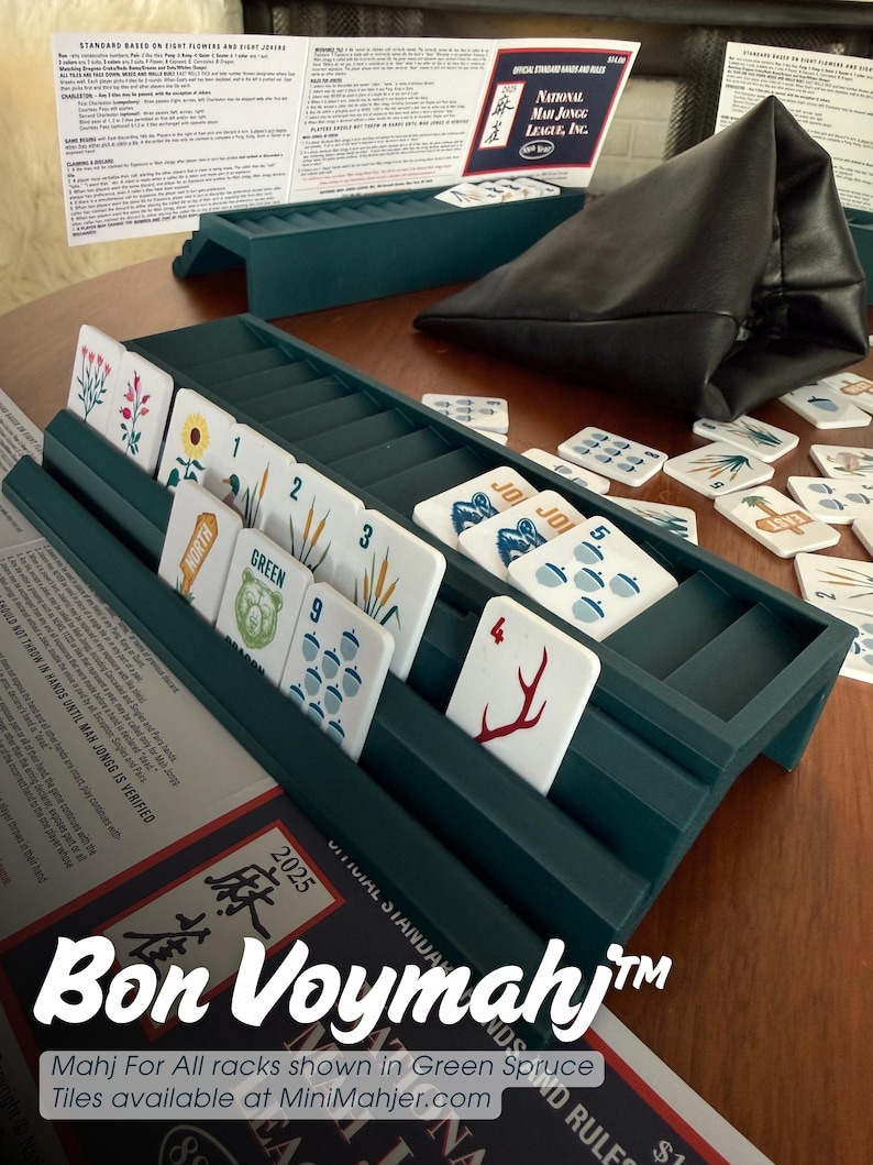 May include: A dark green plastic Mah Jongg rack holding white tiles with various nature-themed illustrations, numbers, and text. A black leather pouch, a printed rule sheet, and additional loose tiles are arranged around the rack on a wooden surface.