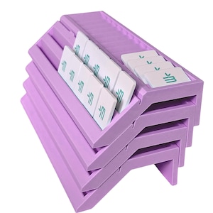 May include: A stack of three lavender-colored plastic organizers with angled slots. Each organizer holds several white rectangular tiles featuring a small green graphic of a stylized plant or leaf icon printed on the surface of each individual tile.