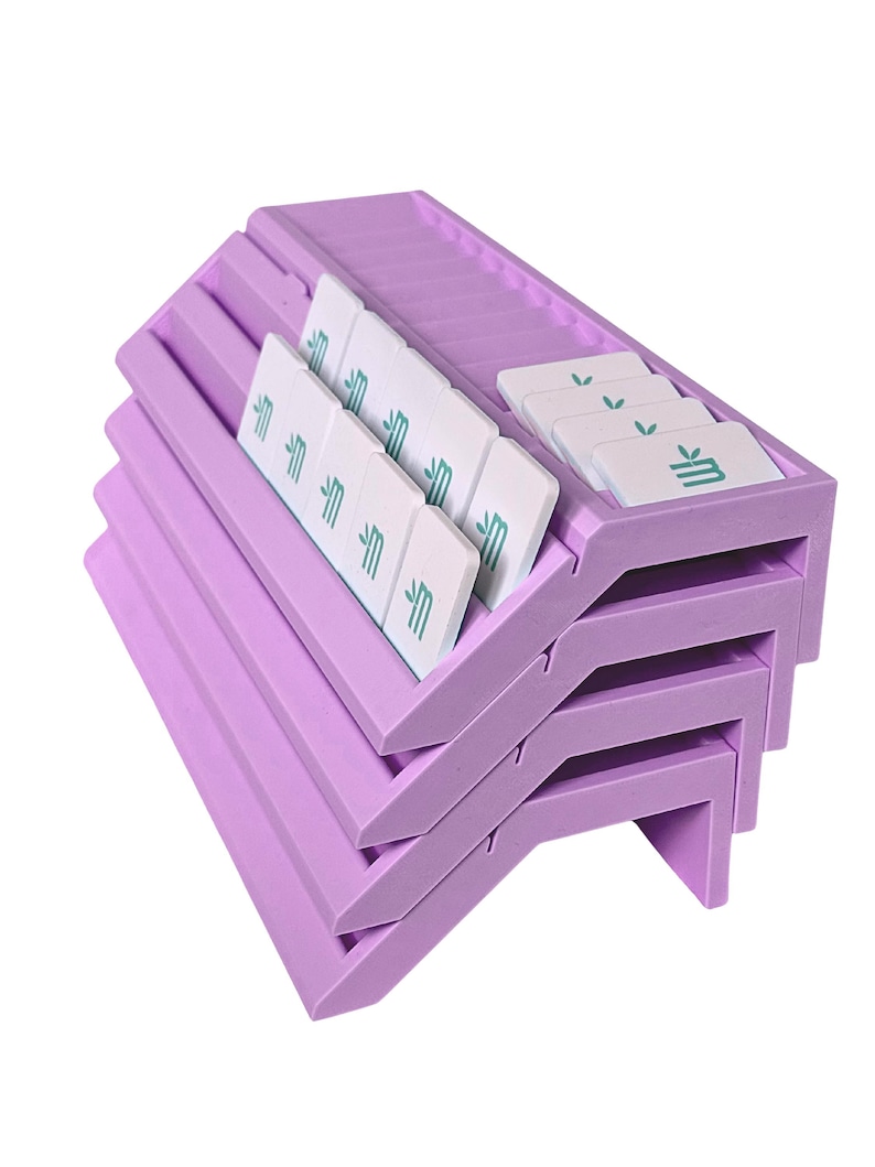 May include: A stack of three lavender-colored plastic organizers with angled slots. Each organizer holds several white rectangular tiles featuring a small green graphic of a stylized plant or leaf icon printed on the surface of each individual tile.