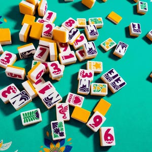 Könnte beinhalten: Eine Ansammlung weißer und gelber Mahjong-Steine, verstreut auf einer türkisfarbenen Oberfläche. Die Steine zeigen rote Zahlen und Symbole, sowie dekorative Muster und das Wort "Joker".