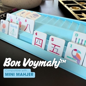 May include: A light blue plastic tile rack holding white game tiles with various colorful illustrations, numbers, and Chinese characters. The text "Bon Voymahj" and "Compatible with... MINI MAHJER" is printed in white and blue on the front of the rack.