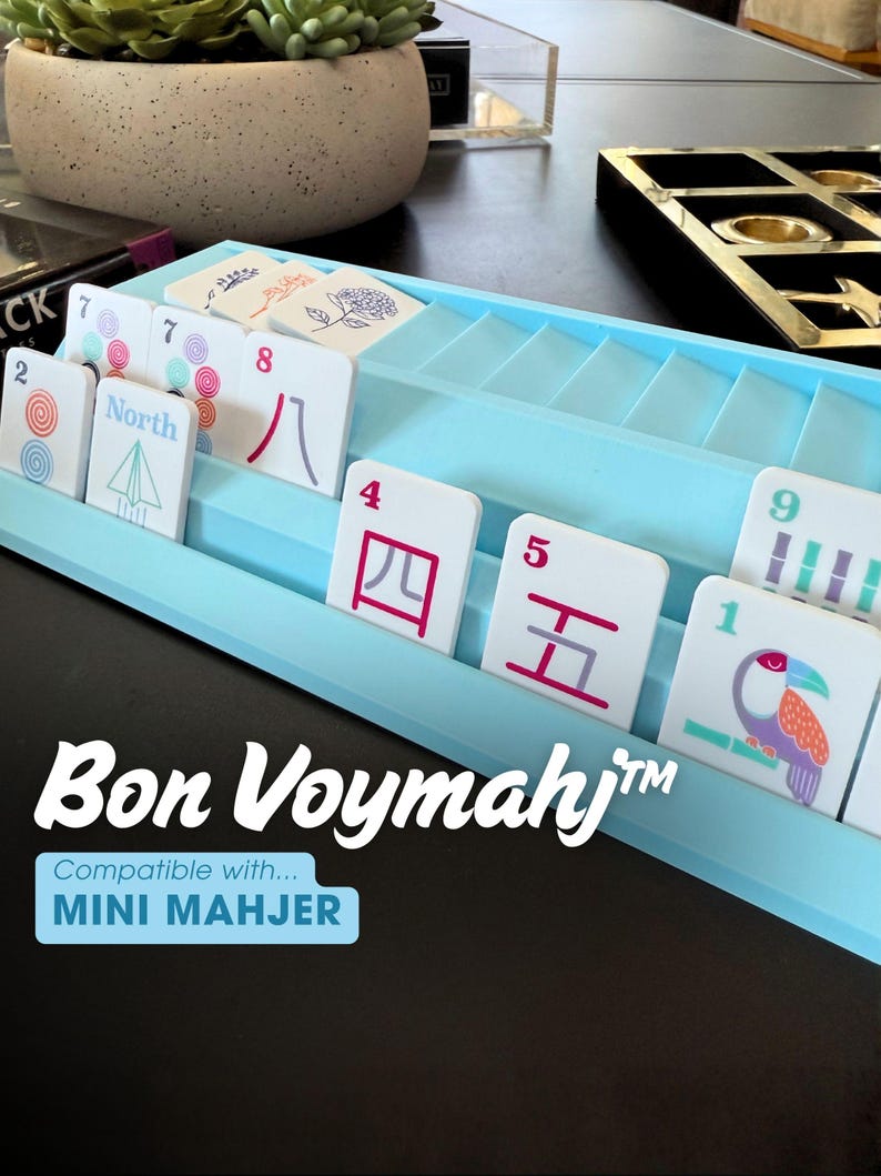May include: A light blue plastic tile rack holding white game tiles with various colorful illustrations, numbers, and Chinese characters. The text "Bon Voymahj" and "Compatible with... MINI MAHJER" is printed in white and blue on the front of the rack.