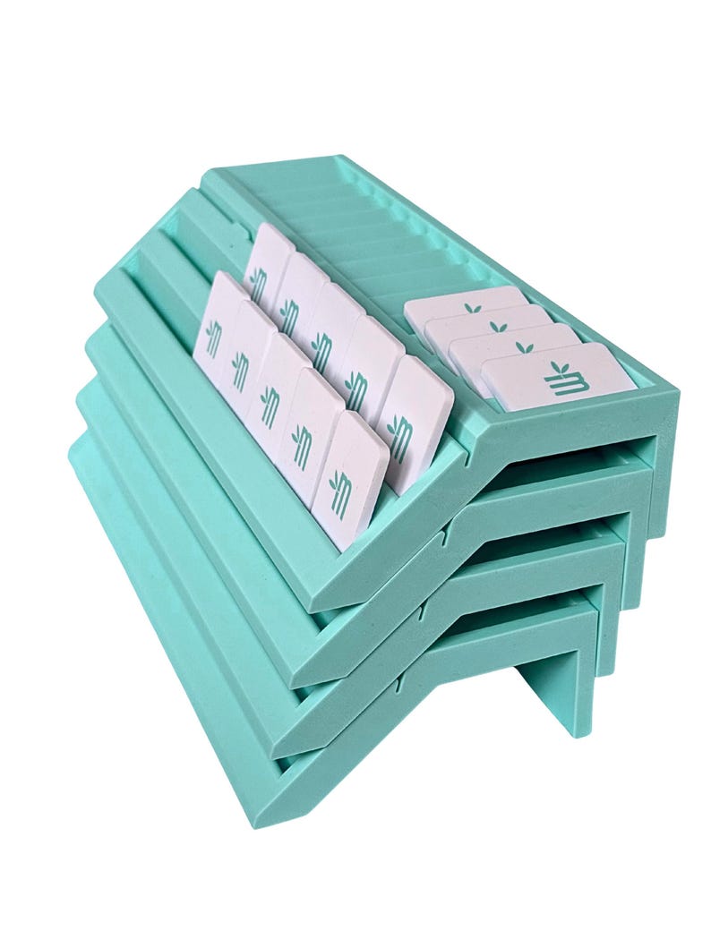 May include: A stack of four light mint green plastic card holders with angled slots. Each holder contains several white rectangular cards featuring a small teal leaf icon and a stylized letter M printed on the front surface of each card.