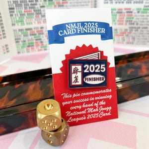 May include: A red and white card with a pin that reads "NMJL 2025 CARD FINISHER" and two gold dice. The card has text that reads "This pin commemorates your success in winning every hand of the National Mah Jongg League's 2025 Card."