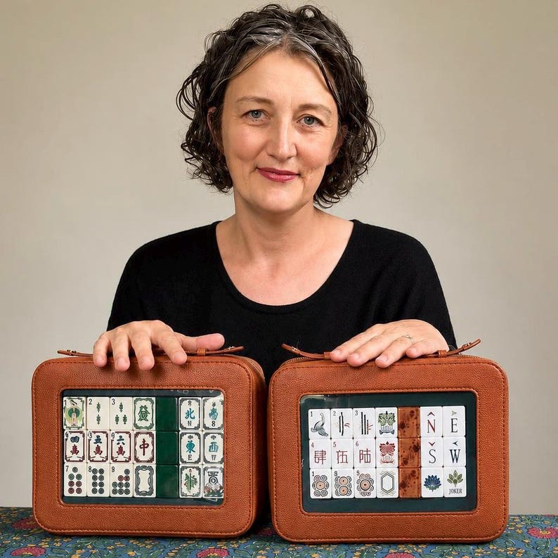 May include: Two brown leather Mahjong tile cases with clear windows displaying the tiles. The tiles are white with black and green symbols. The cases are rectangular with a zipper closure. The woman in the image is wearing a black shirt.