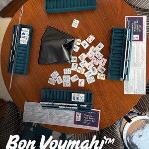 May include: A Mah Jongg game set featuring four teal plastic tile racks, a black storage pouch, and a scattered collection of white tiles with colorful symbols. Two National Mah Jongg League rule cards are placed on the round wooden table.
