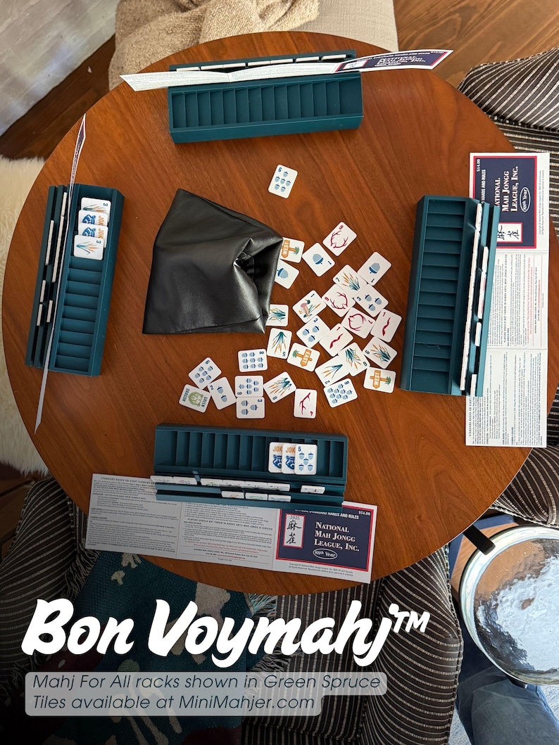 May include: A Mah Jongg game set featuring four teal plastic tile racks, a black storage pouch, and a scattered collection of white tiles with colorful symbols. Two National Mah Jongg League rule cards are placed on the round wooden table.
