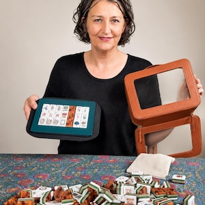 Op de afbeelding: Een vrouw toont een Mahjong-set, bestaande uit tegels, een zwarte en teal draagtas en een bruine etui met ritssluiting. De tegels zijn bruin, wit en groen. Het spel wordt gepresenteerd op een oppervlak met bloemenpatroon.