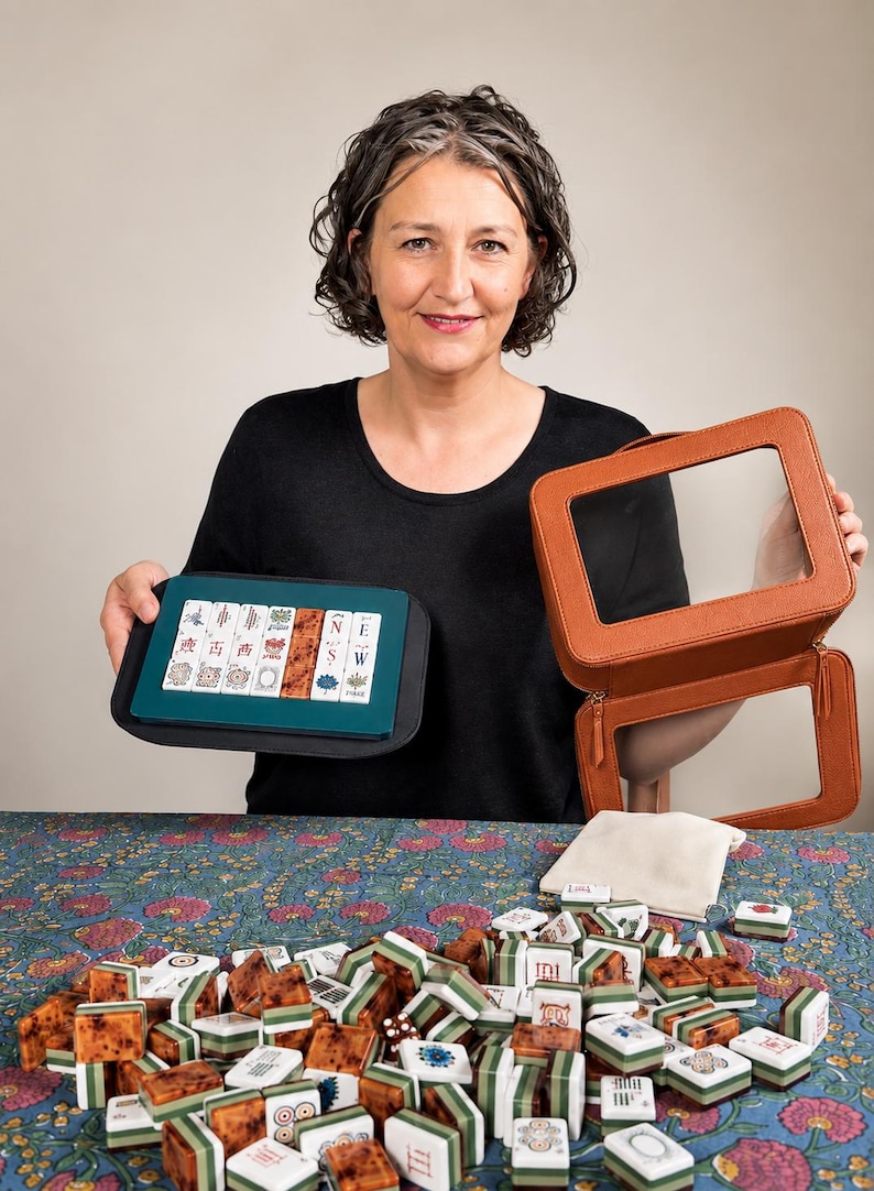 May include: A woman holds a Mahjong set, including tiles, a carrying case, and a zippered case. The tiles are brown, white, and green. The carrying case is teal and black, and the zippered case is brown. The game is displayed on a floral patterned surface.