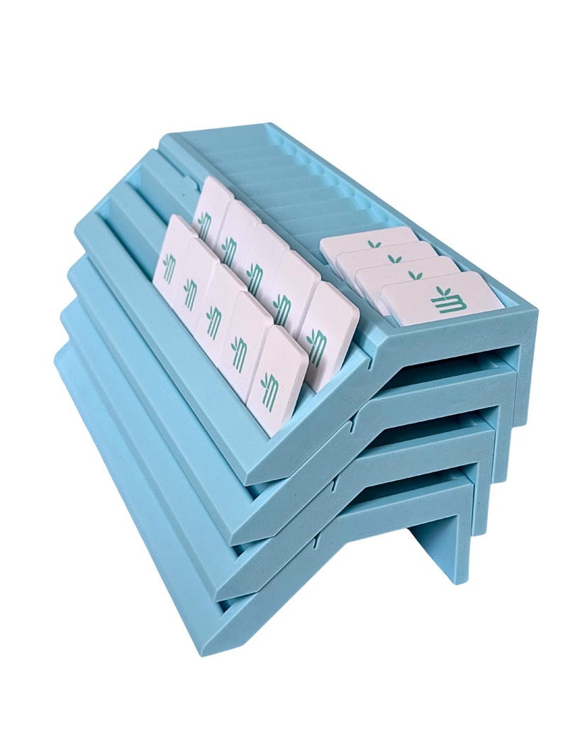 May include: A stack of four light blue plastic card holders with angled slots. Each holder features white rectangular tiles with a green leaf icon and a stylized letter M printed on them, arranged in the grooves of the top holder.