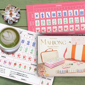 Può includere: Una composizione di natura morta con un set di Mahjong, una tazza di matcha latte e una pianta succulenta su una superficie di legno verde. Il set di Mahjong include tessere, una custodia e una borsa a righe. Sono visibili anche grafici di gioco rosa e bianchi.