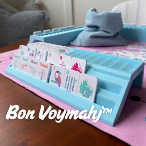 May include: A light blue plastic tile rack holding white game tiles with colorful illustrations of paper airplanes, a toucan, bamboo, and a dragon. The tiles feature text including North, East, West, South, and the brand name Bon Voymahj.