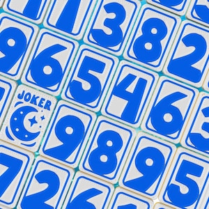 May include: Close-up of a game tile set with blue numbers and a "JOKER" card. The tiles have a white background with blue borders and numbers. The "JOKER" card features a moon and star design.