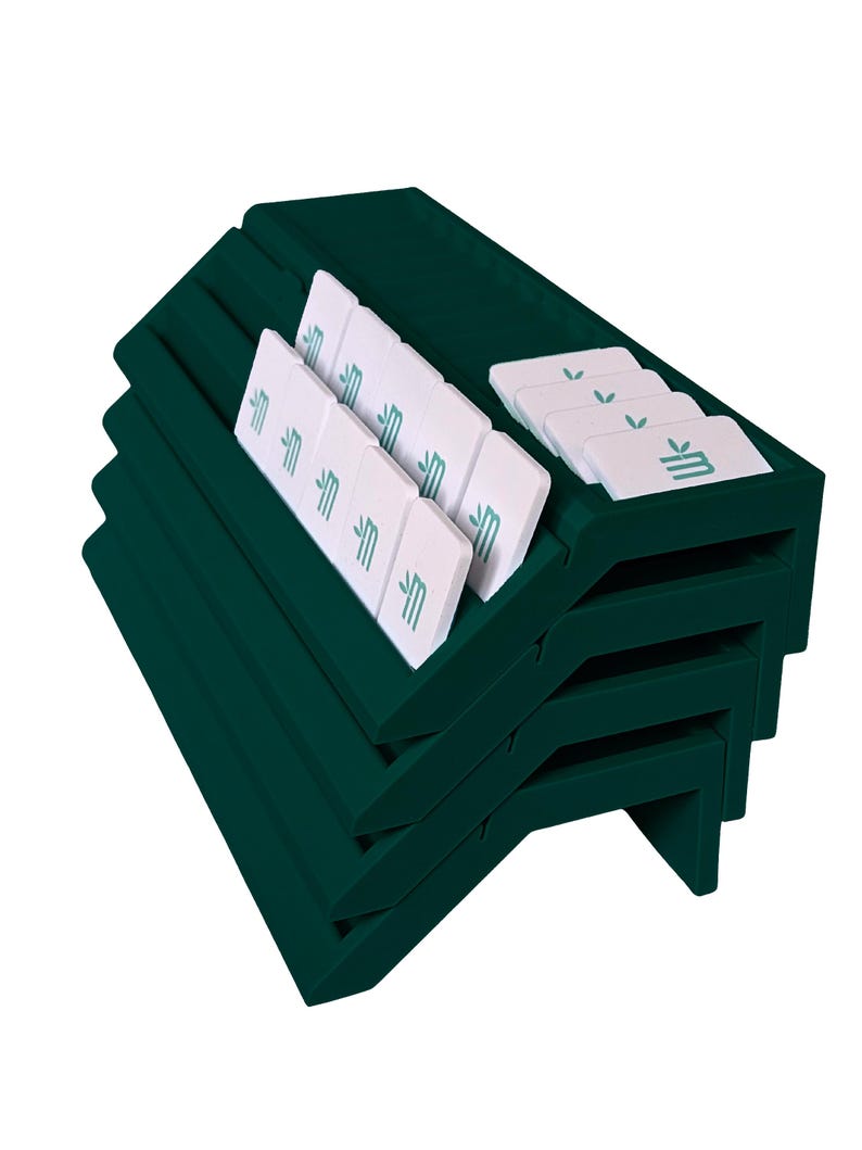 May include: A dark green tiered display rack featuring multiple angled slots. Several rectangular white tiles are arranged within the slots, each printed with a teal logo consisting of a stylized letter M and two small leaves above it.