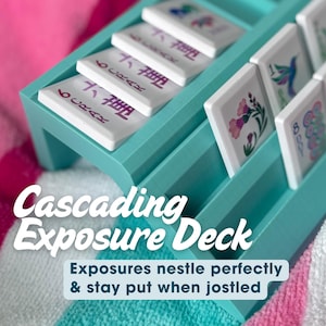 May include: A teal plastic cascading exposure deck rack for mahjong tiles. The rack features tiered slots holding white tiles with colorful floral and numerical designs. Text on the image reads: Cascading Exposure Deck, Exposures nestle perfectly & stay put when jostled, Shown at Gulf Shores, with travel tiles from OhMyMahjong.com.