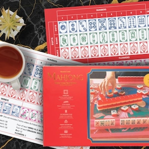 May include: A red Mahjong game set with a gold-colored title and a detailed illustration of the game in progress. The set includes tiles, a game board, and instructions. A cup of tea and a small plant are also visible.