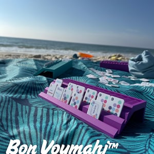 May include: A purple plastic rack holding white game tiles with colorful circular patterns and text including Joker, West, and Soap. A teal rack sits nearby. The text Bon Voymahj and Mahj For All racks shown in Purple Orchid & Aqua Tiles available at MiniMahjer.com is visible.