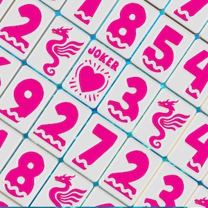 May include: Close-up of a collection of white game tiles with bright pink numbers, dragon illustrations, and the word "JOKER" with a heart symbol. The tiles are arranged in a grid pattern with a light blue background.