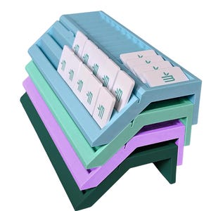 May include: A stack of four angled, rectangular organizers in light blue, mint green, lavender, and dark green. The top light blue organizer features multiple slots holding white rectangular tiles, each printed with a small green leaf icon.