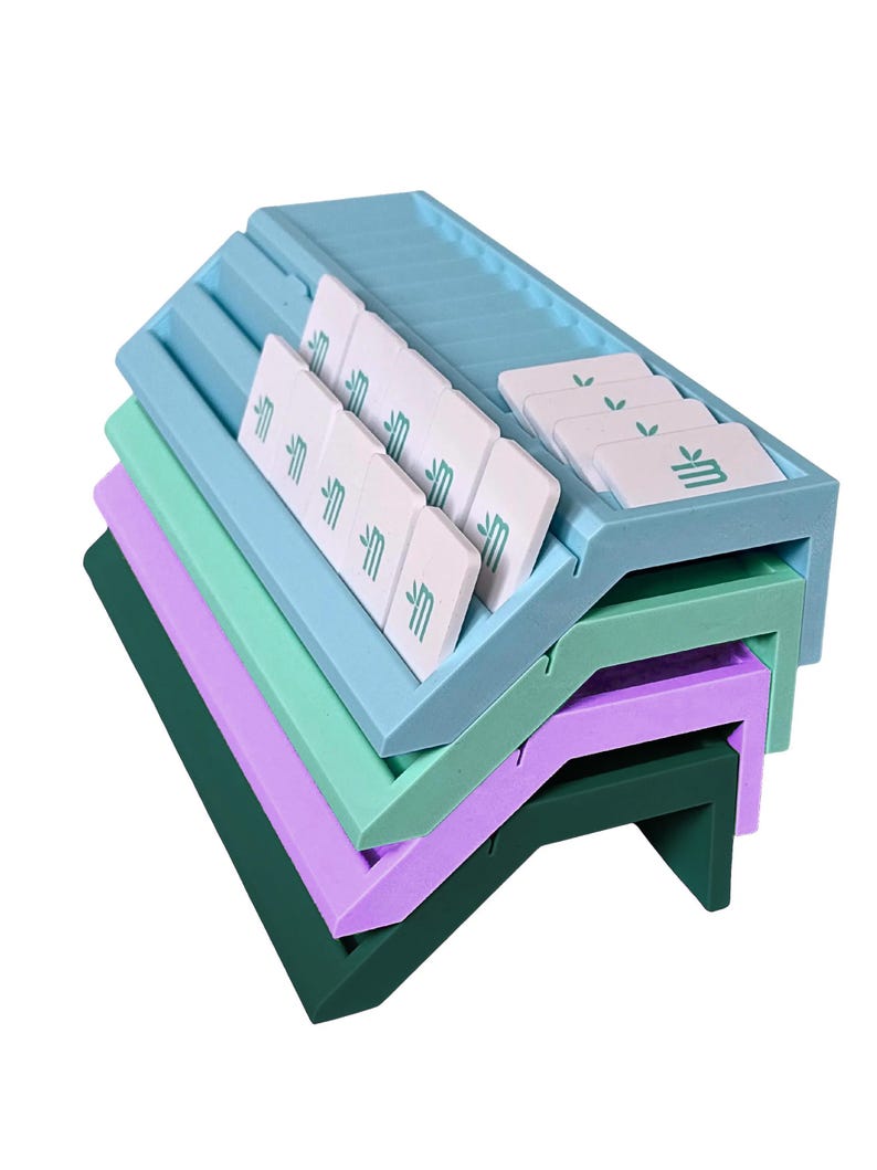May include: A stack of four angled, rectangular organizers in light blue, mint green, lavender, and dark green. The top light blue organizer features multiple slots holding white rectangular tiles, each printed with a small green leaf icon.