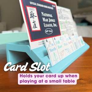 May include: A light blue plastic card holder stand designed to support a National Mah Jongg League 2025 official standard hands and rules card. The stand features a grooved slot for upright card placement, shown with game tiles.