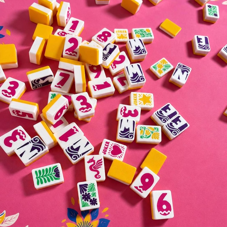 May include: A selection of colourful Mahjong tiles scattered on a pink surface. The tiles display numbers, symbols, and characters in various colours, including red, purple, green, and yellow. Some tiles have the word "Joker" printed on them.