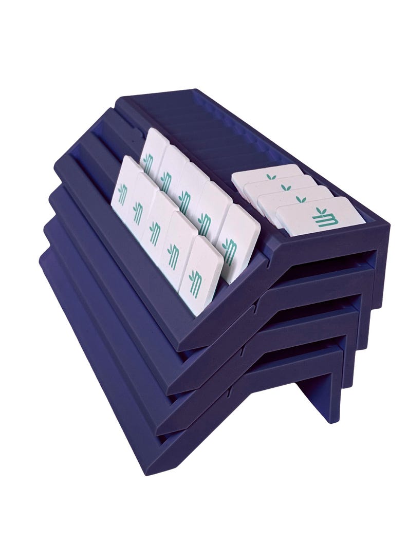 May include: A dark blue tiered display rack with multiple horizontal slots holding several white rectangular tiles. Each tile features a small green graphic of a stylized letter M with a leaf icon above it, arranged in organized rows.