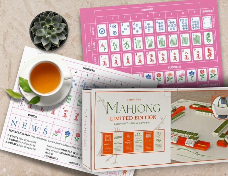 K&ouml;nnte beinhalten: Ein Stillleben mit einem Mahjong-Set in limitierter Auflage mit einem rosa Spielbrett, Spielsteinen und einer Tasse Tee. Auf der Box steht "Mahjong Limited Edition". Das Spielset enth&auml;lt Spielsteine und ein rosa Spielbrett mit Zahlen und Symbolen.