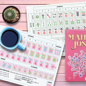 May include: A pink Mah Jong game box with the words "MAH JONG" in gold lettering, game tiles, and game instructions. A blue cup of coffee and a small cactus in a black pot are also in the image.
