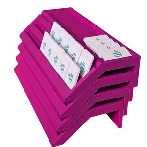 May include: A stack of four bright magenta plastic card holders with angled slots. Each holder features multiple grooves designed to display small white rectangular cards. The cards are printed with a teal logo featuring a stylized letter M.