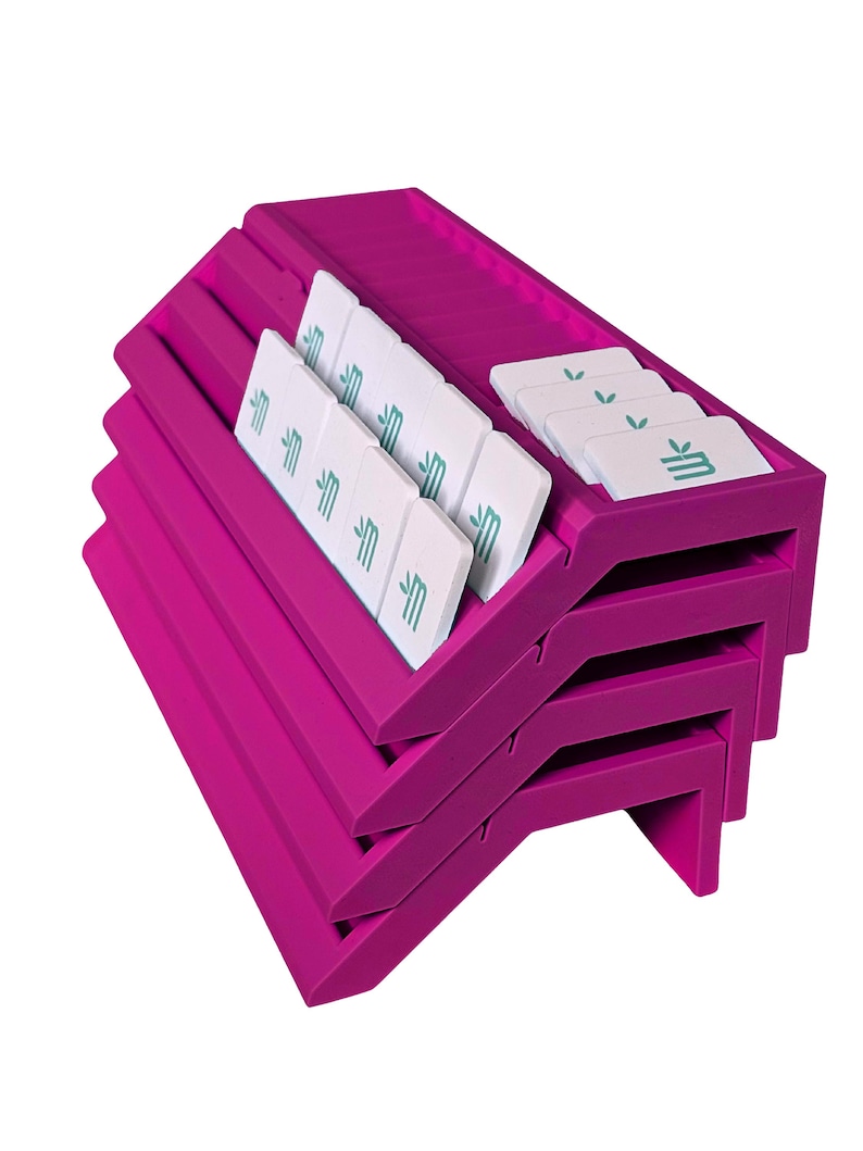 May include: A stack of four bright magenta plastic card holders with angled slots. Each holder features multiple grooves designed to display small white rectangular cards. The cards are printed with a teal logo featuring a stylized letter M.