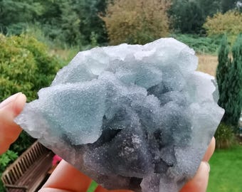 Puede incluir: Una mano sostiene un grupo de cristales de fluorita azules y grises. La formación mineral tiene una superficie texturizada y cristalina con diferentes tonos de azul y gris. El fondo muestra follaje verde y un banco de madera.