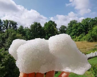 Può includere: Una mano tiene in mano un grande esemplare di minerale cristallino bianco contro un cielo blu e alberi verdi. Il minerale ha una superficie ruvida e strutturata e una forma a nuvola. Lo sfondo comprende un prato verde e una recinzione bianca.