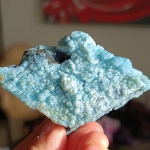 May include: A close-up of a light blue mineral specimen with a bubbly, textured surface. The mineral has a crystalline structure and is held in a hand. The background is blurred, with hints of red and neutral tones.