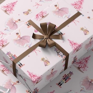 Puede incluir: Una caja de regalo envuelta con una cinta y un lazo marrón se encuentra sobre papel de regalo. El papel presenta un patrón repetido de bailarinas rosas, árboles de Navidad y cascanueces. Tijeras y cinta adhesiva en primer plano.