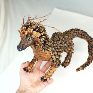 May include: A small dragon sculpture made entirely of pine cones. The dragon has a long tail, a spiky mane, and yellow eyes.