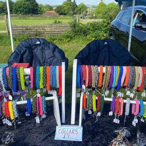 Puede incluir: Un expositor de collares para perros de paracord de colores a la venta en un mercado al aire libre. Los collares están dispuestos en soportes blancos y se venden a 7 € cada uno. El cartel dice "COLLARS 7 €".
