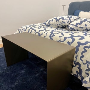 Puede incluir: Una mesa rectangular larga y estrecha hecha de metal con un acabado marrón oscuro. La mesa está sobre una alfombra azul de pelo largo delante de una cama con ropa de cama blanca y azul.