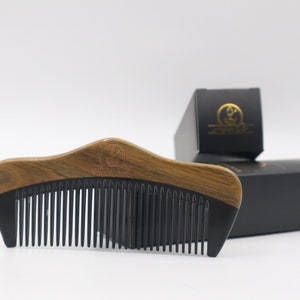 May include: A wooden comb with a dark brown handle and black teeth. The comb has a curved design and a logo on the handle. Two black boxes with gold logos are in the background.