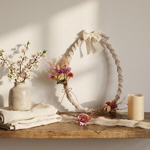 Op de afbeelding: Een decoratieve krans gemaakt van geweven touw, versierd met gedroogde bloemen en een crèmekleurige strik. Een kleine vaas met witte bloemen, gevouwen linnen en een kaars staan op een houten plank. Een kanten lint en een klein bloemstuk zijn ook aanwezig.
