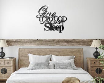 May include: Black metal wall decor with the text "Give it to God and go to sleep". The decor is hanging above a wooden headboard with white bedding.