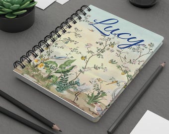 May include: A spiral bound notebook with a floral design and the name "Lucy" printed in blue on the front cover.